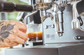 A tattooed hand holding a graduated shot glass under a commercial espresso machine group head