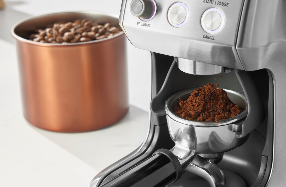 Fine espresso grind dosing into a portafilter from a burr grinder beside copper bean canister