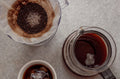 An overhead view of a pour over dripper with used coffee grounds next to a glass server and espresso cup