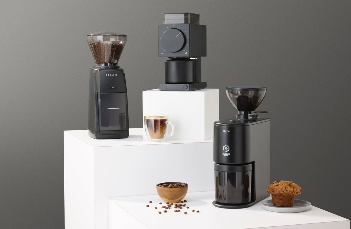Baratza, Fellow Ode, and Eureka Peppe coffee grinders on white display blocks with coffee
