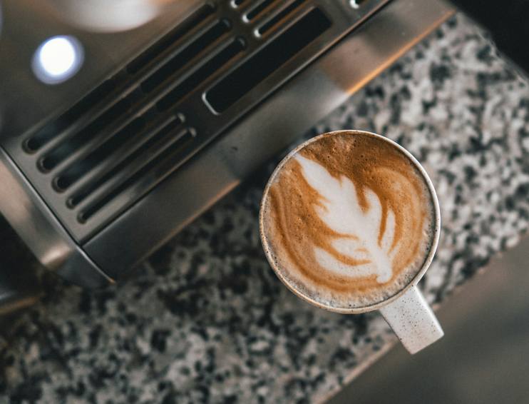 A cappuccino with latte art in a small ceramic cup sitting on a granite countertop next to an espresso machine - Seattle Coffee Gear