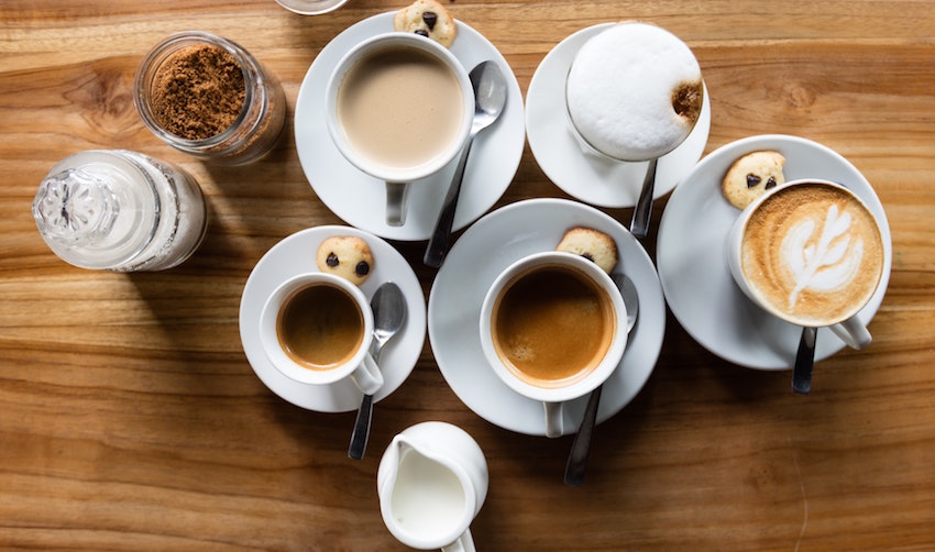 A flat lay of coffee drinks including lattes and espresso alongside a sugar bowl, milk jug, and spoons arranged on a table - Seattle Coffee Gear