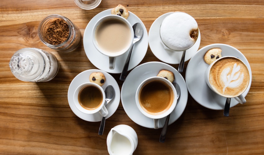 A flat lay of coffee drinks including lattes and espresso alongside a sugar bowl, milk jug, and spoons arranged on a table - Seattle Coffee Gear