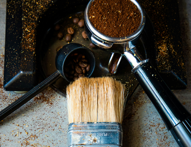 Espresso machine cleaning tools including a group head brush, portafilter with coffee grounds, and a knock box on a countertop
